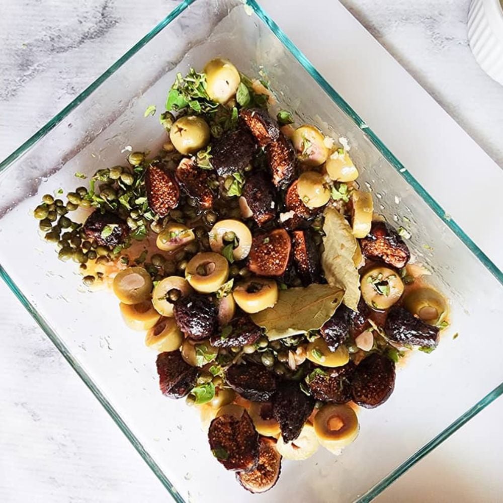 a picture of chicken marbella marinating with olives figs and capers in a glass dish