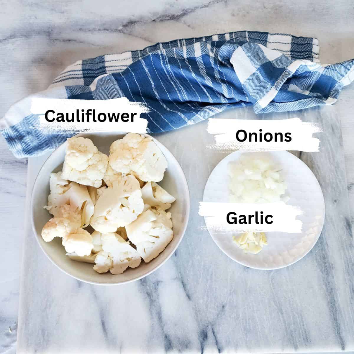 overhead image of the ingredients to make cauliflower soup with cut cauliflower in a bowl, onions and garlic