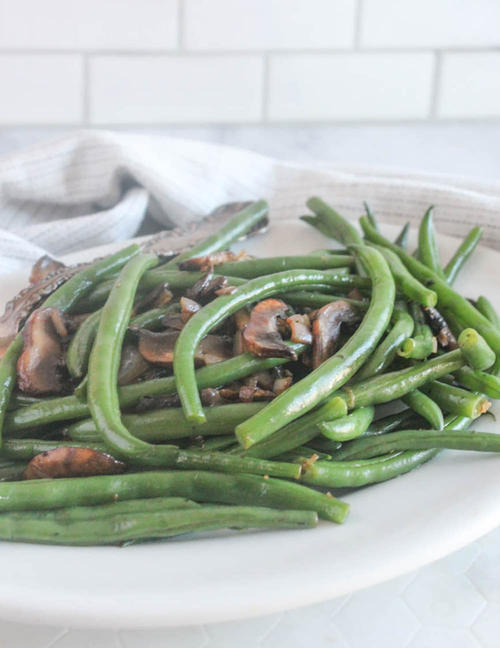 A close up picture of sauteed green beans with mushrooms on a white serving plate