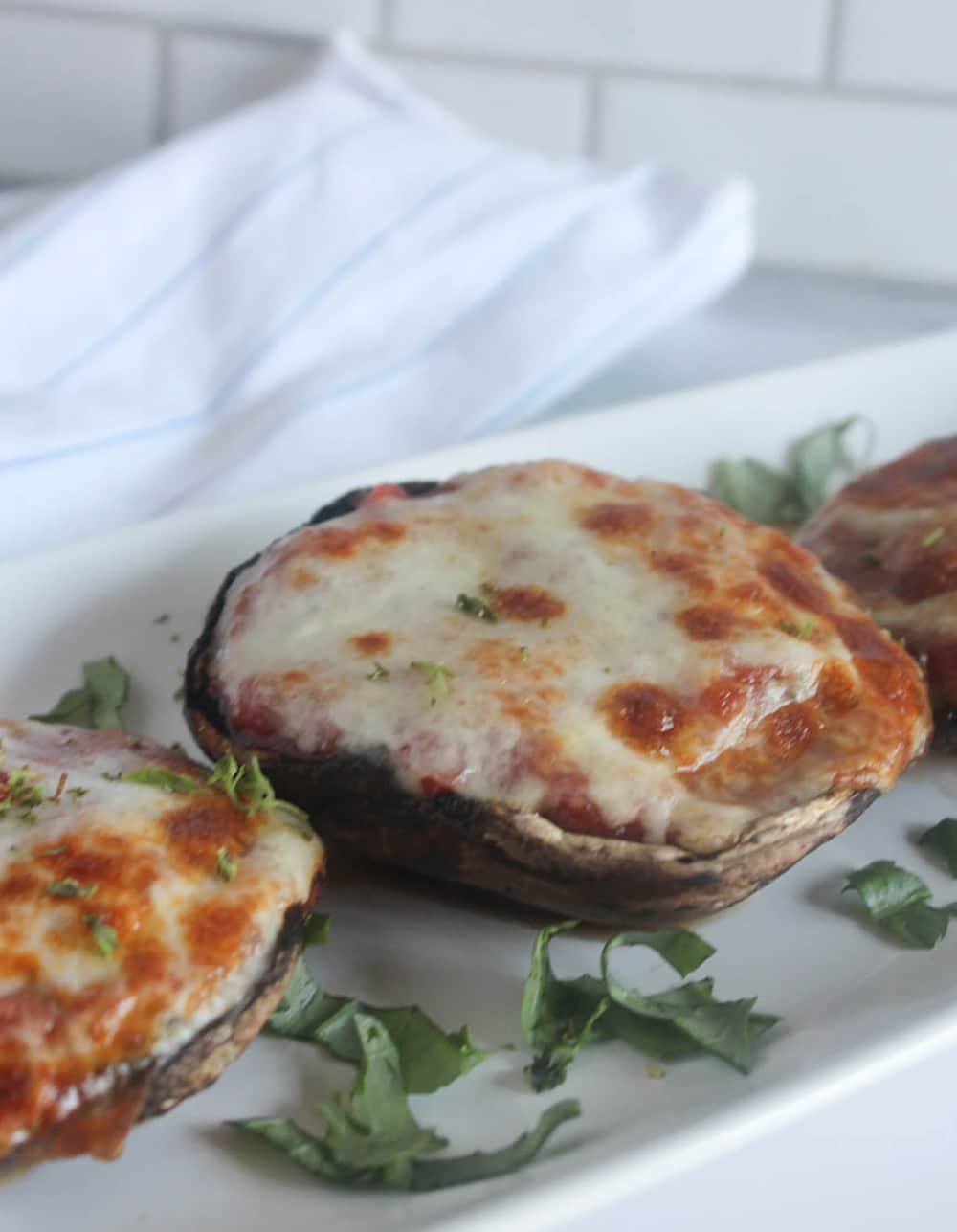This image shows three portobello mushroom caps placed on an oblong white tray with pieces of fresh basil scattered on the tray
