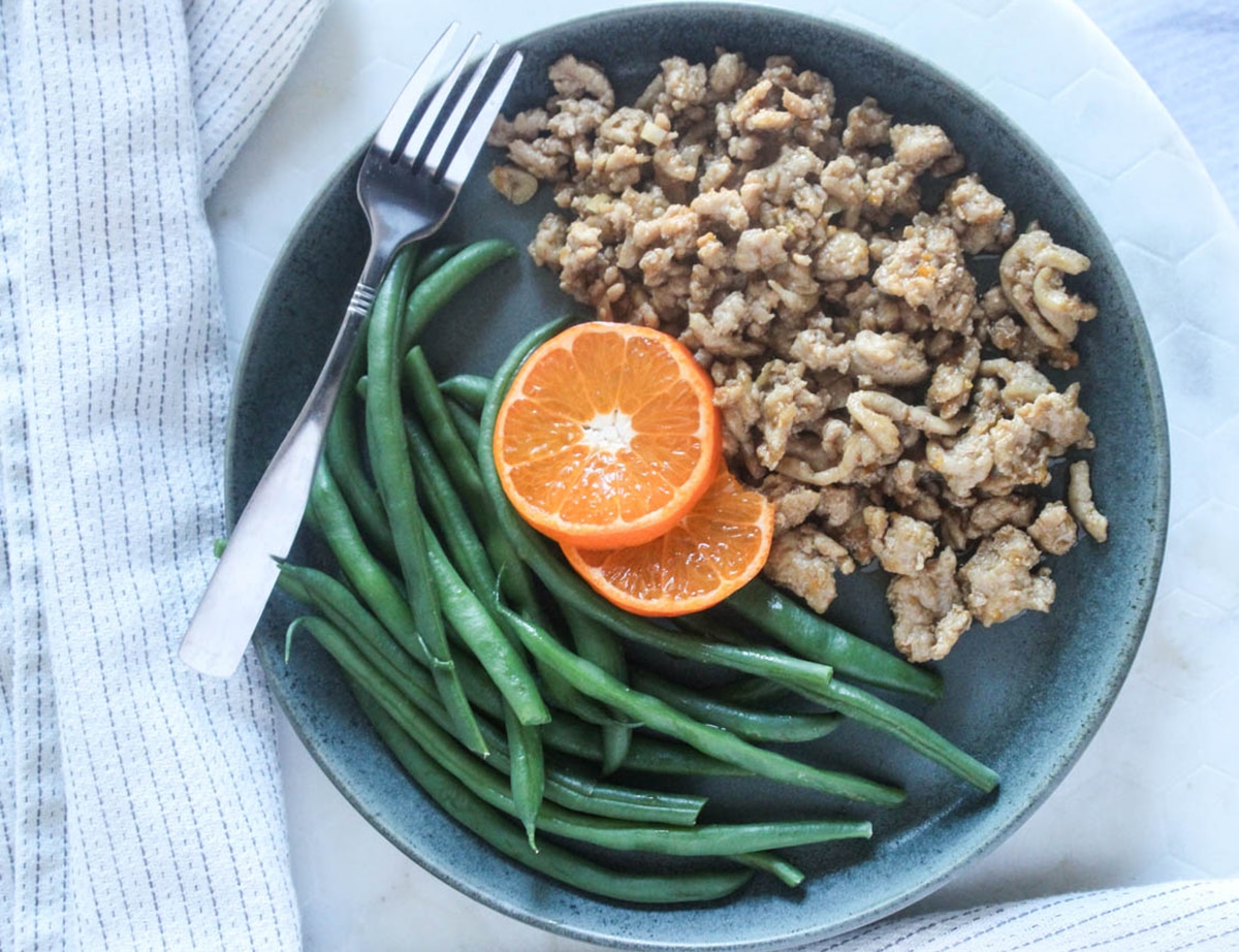 An overhead shot of ground orange chicken on a plate with green beans and orange slices