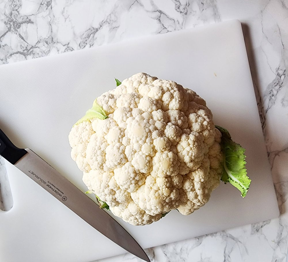 A picture of a head of cauliflower on a cutting board with a knife.