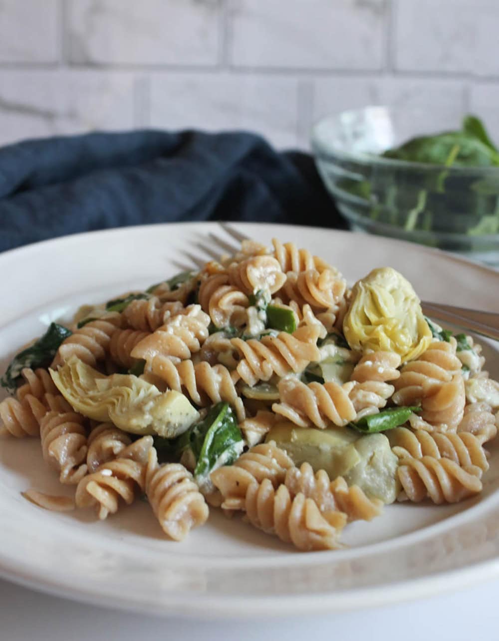 a close up picture of spinach artichoke pasta on a plate with a fork and a bowl of salad in the background