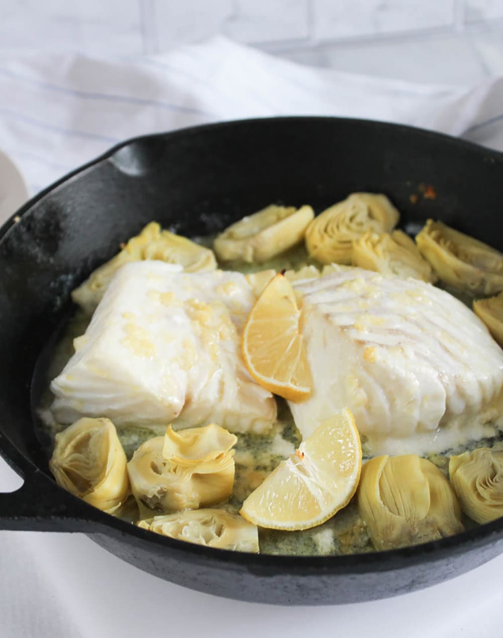 a picture of two codfish filets with artichokes, lemons in a heavy cast iron skillet