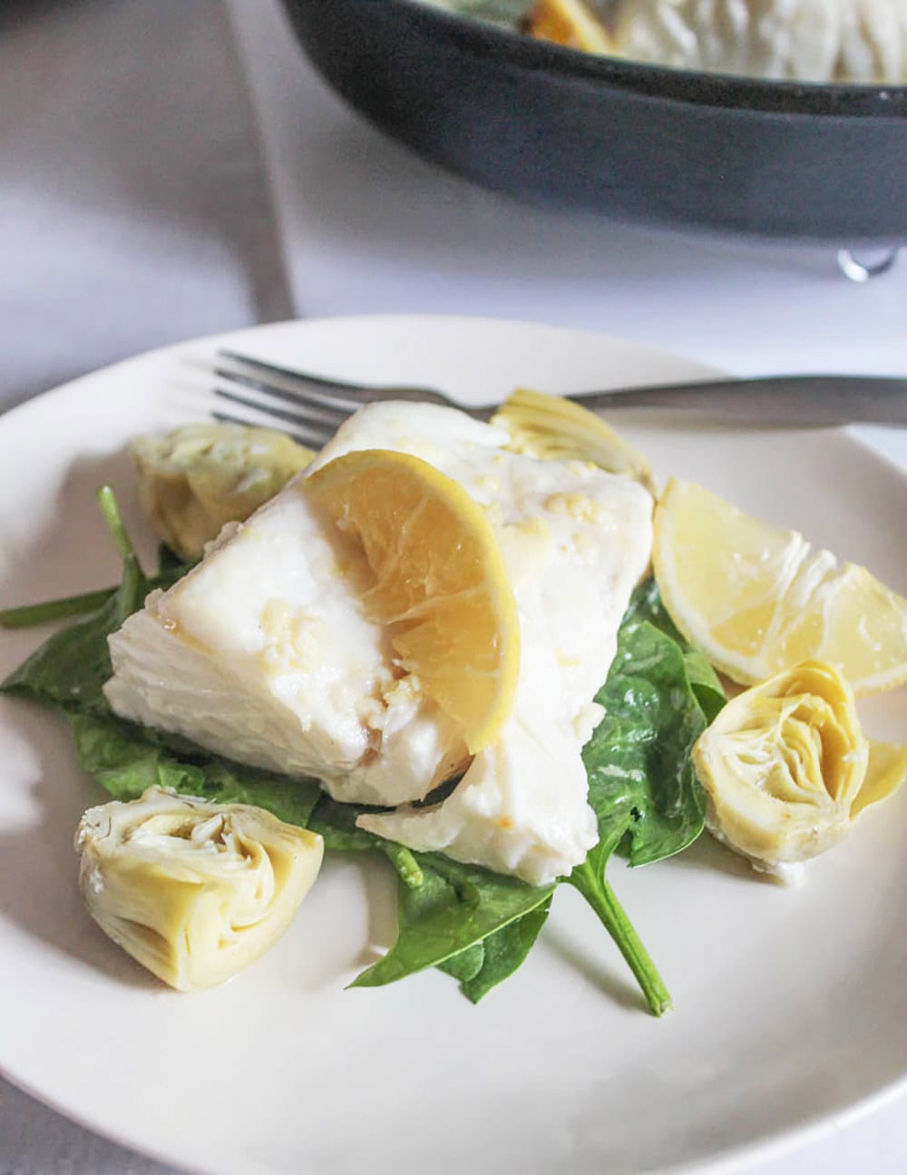 a codfish filet on top of spinach on a white plate with artichoke hearts and lemon