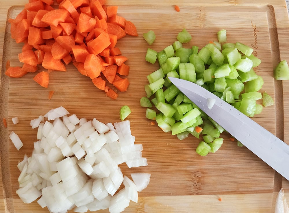 Mirepoix process step 1: a picture of chopped onion, carrots and celery on a cutting board with a knife