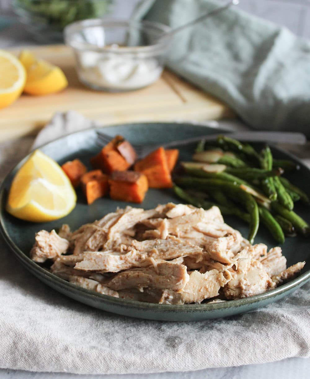 cooked and shredded greek yogurt chicken on a plate with green beans, sweet potatoes and a wedge of lemon