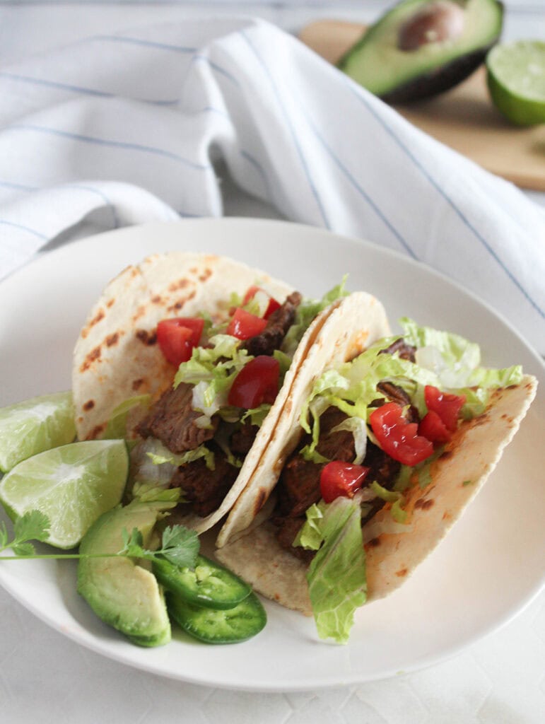 two shredded beef tacos on a white plate with lime wedges and jalapeno slices