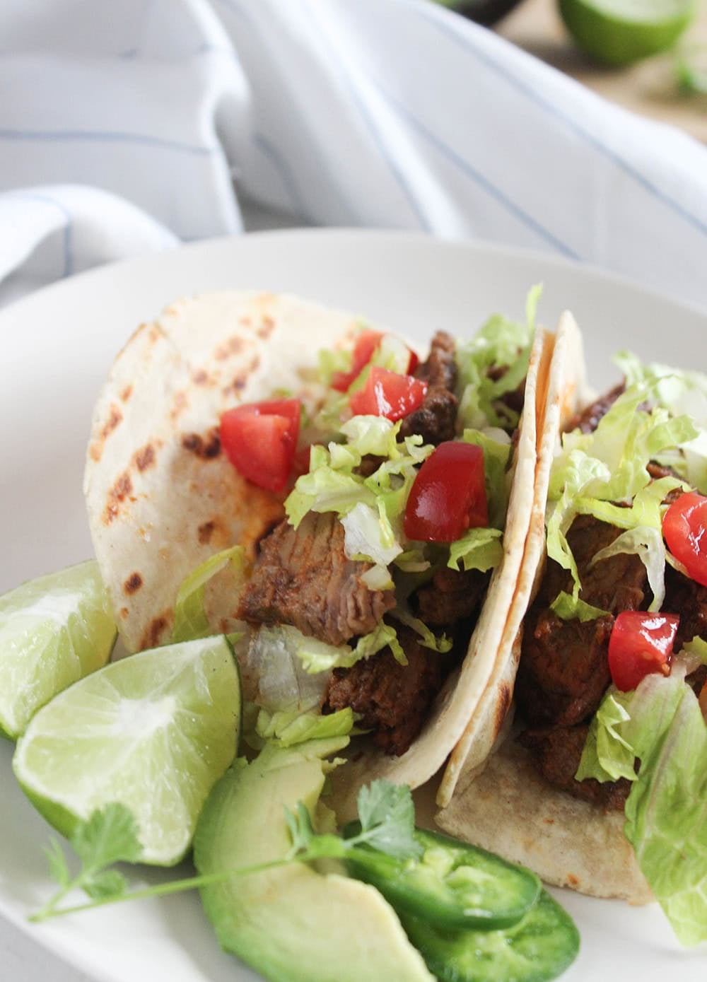 a picture of two slowcooker beef tacos on a white plate topped with shreds of lettuce, tomatoes with lime wedges on the side