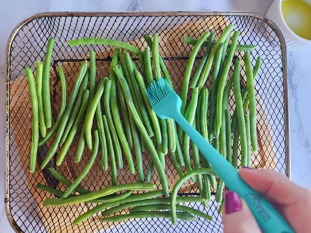 green beans arranged in an air fryer basket witih a pastry brush brushing on oil