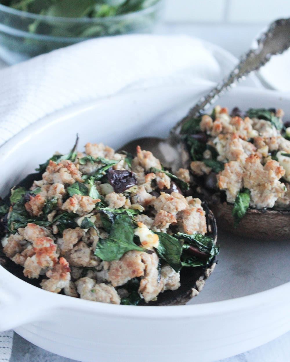 Close-up of baked portobello mushrooms stuffed with a savory mixture of ground turkey, wilted spinach, and crumbled cheese, served in a white ceramic dish with tongs resting on the side.
