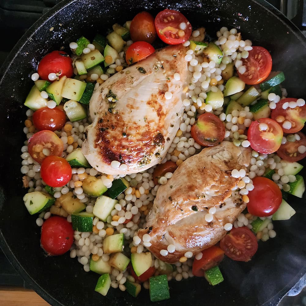 a picture of the finished dish. two chicken breasts surrounded by couscous, cherry tomatoes and zucchini tossed in pesto sauce