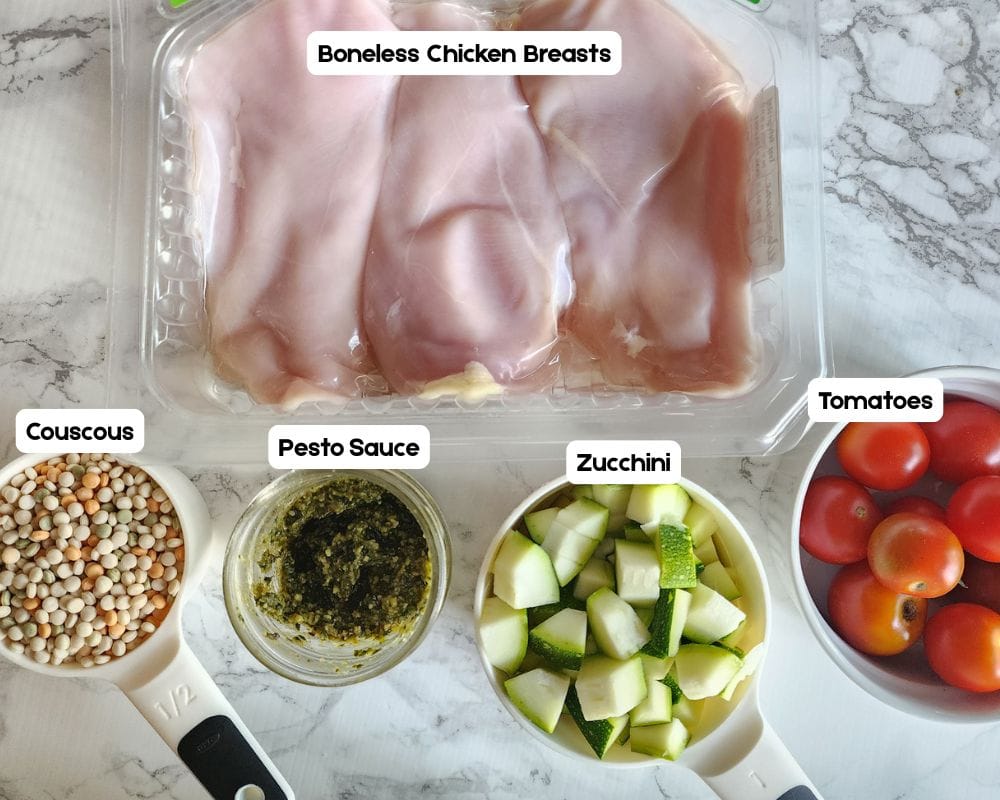 a view of the ingredients needed to make this chicken pesto recipe: couscous, pesto sauce, zucchini, cherry tomatoes and boneless chicken breasts