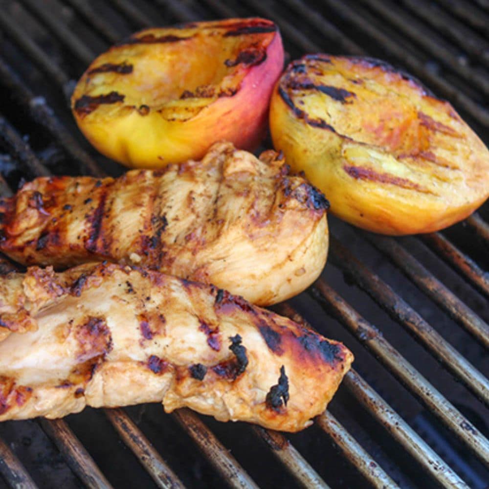 a picture showing two boneless chicken breasts and a peach cut in half cooking on the grill.