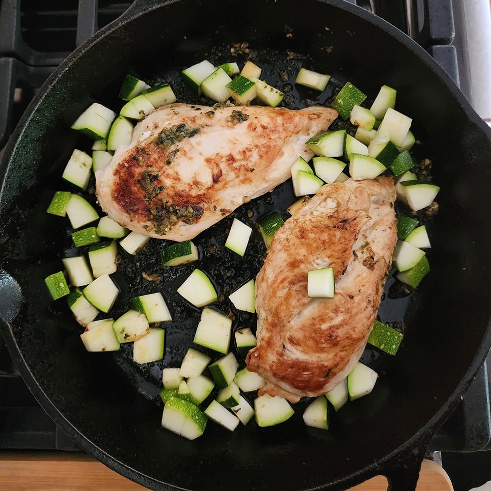 two chicken breasts cooking in a skillet with zucchini cubes and pasto sauce 
