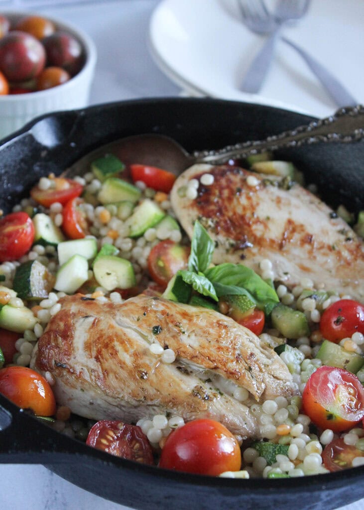 two cooked boneless pesto chicken breasts in a cast iron skillet with zucchini cubes, cherry tomato halves and couscous