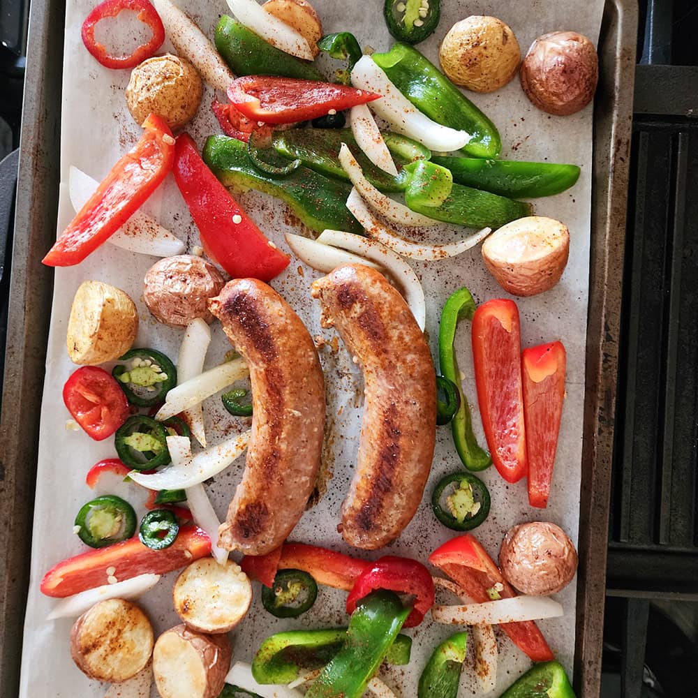 an overhead view process step 3. Cooked sausages and potatoes on a sheet pan with uncooked peppers and onions