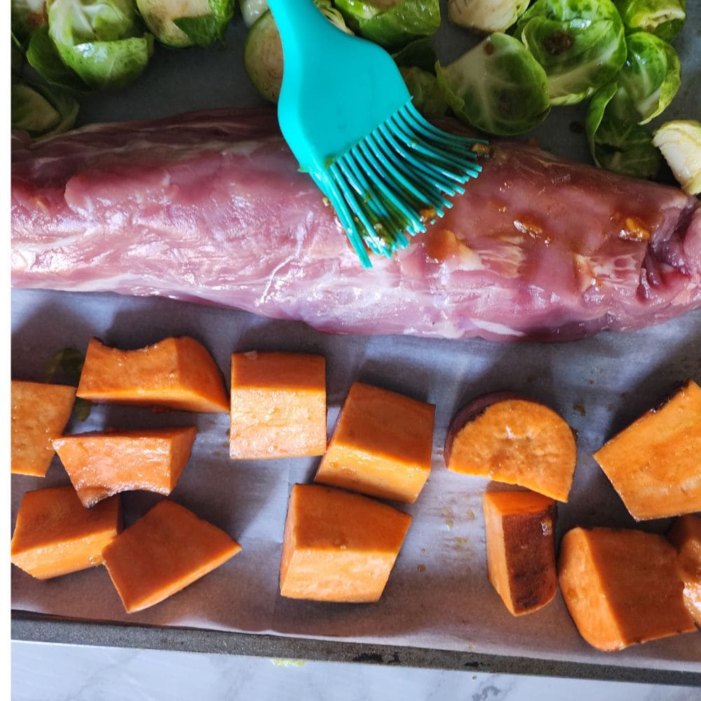 a picture of pork tenderloin on a sheet pan with sweet potatoes with sauce brushing on.