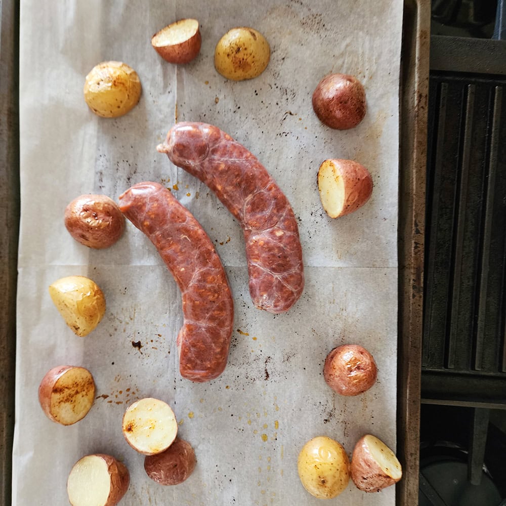 two uncooked sausages on a sheet pan with cooked sliced potatoese