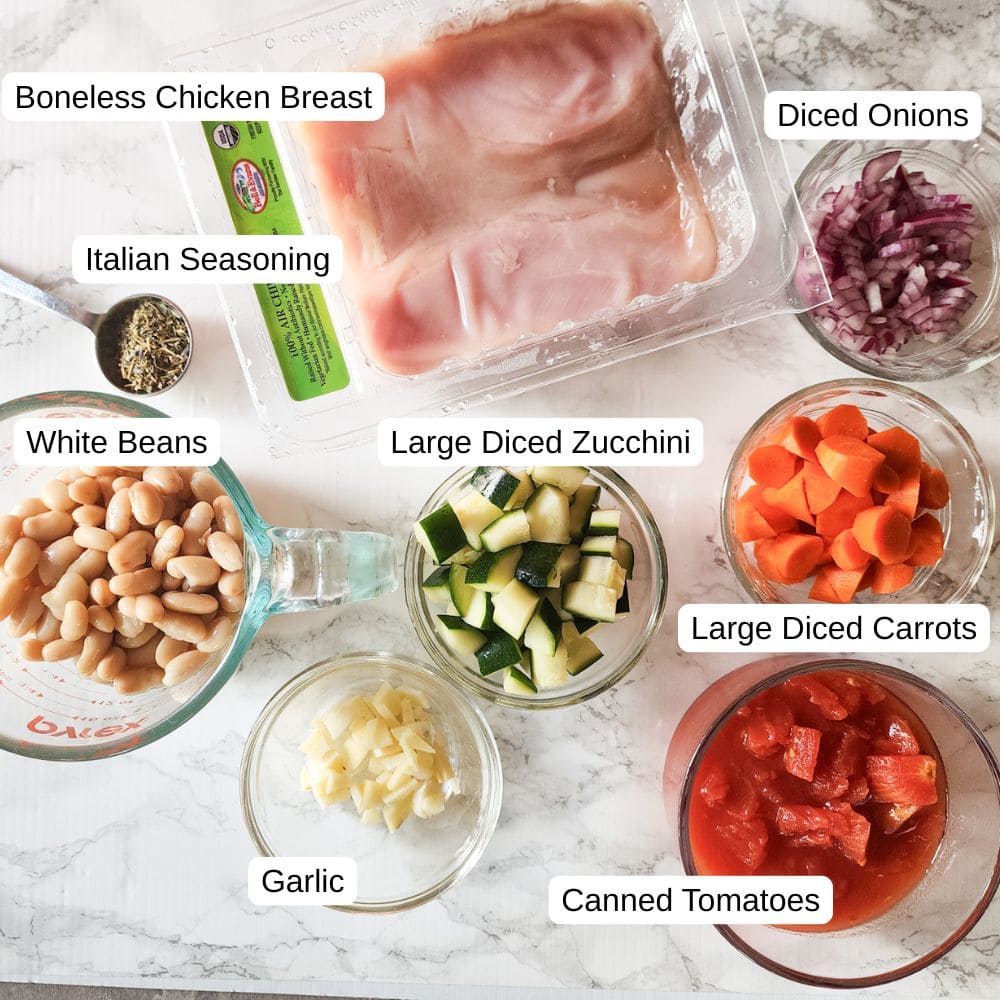 the ingredients for tuscan chicken laid out on a marble board: white beans, Italian seasoning, Chicken breasts, onions, carrots, zucchini, garlic, onions and tomatoes