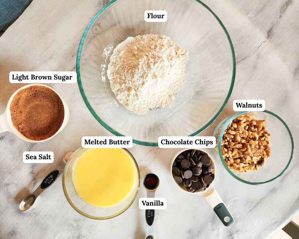 a picture of the ingredients needed to make chocolate chip cookies brittle: flour, brown sugar, salt, melted butter, vanilla, chocolate chips, walnuts