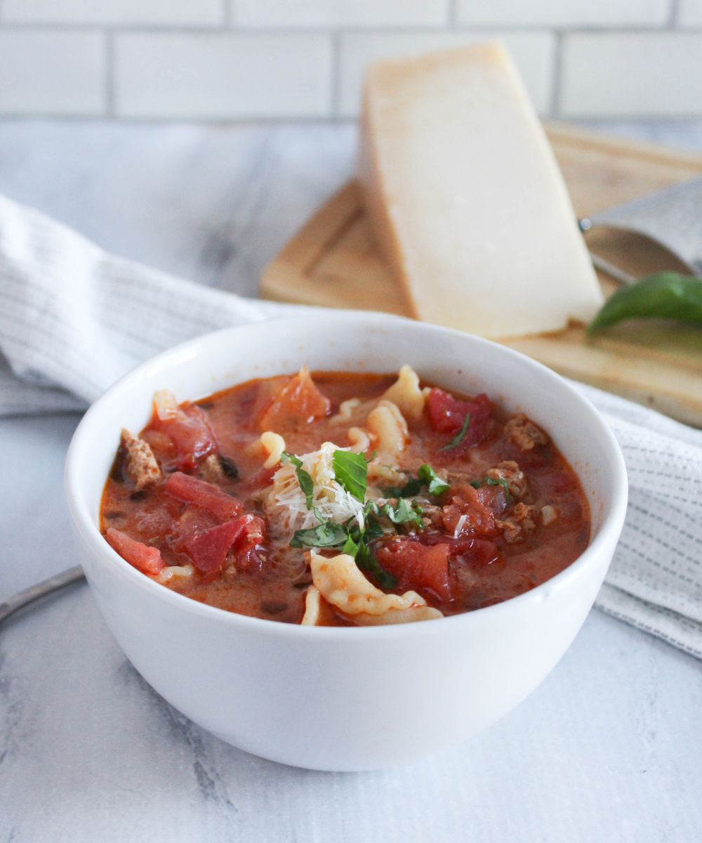 lasagna soup in a white soup bowl with a wedge of parmesan cheese on a cutting board behind it