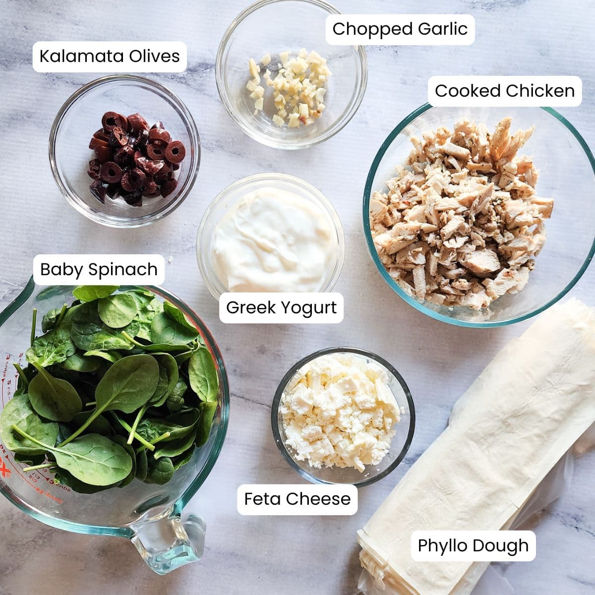 a photograph of the ingredients needed to make greek chicken casserole: spinach, olives, chicken, phyllo dough, greek yogurt, garlic and feta cheese