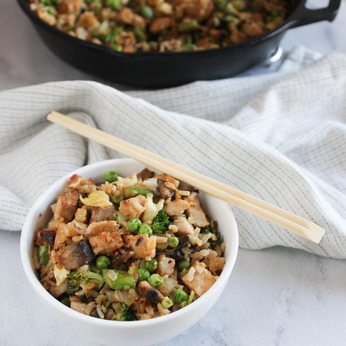 a picture of fried rice in a white bowl with chopsticks on the side and a skillet with  fried rice in the background